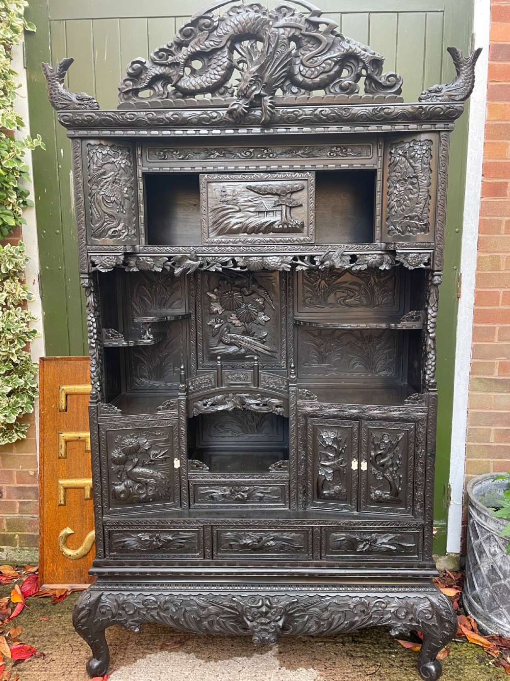 An early 20th-century Japanese ebonised timber shodana display cabinet featuring detailed dragon and floral relief carvings, asymmetrical shelving, small cupboard doors, and a carved stand base.