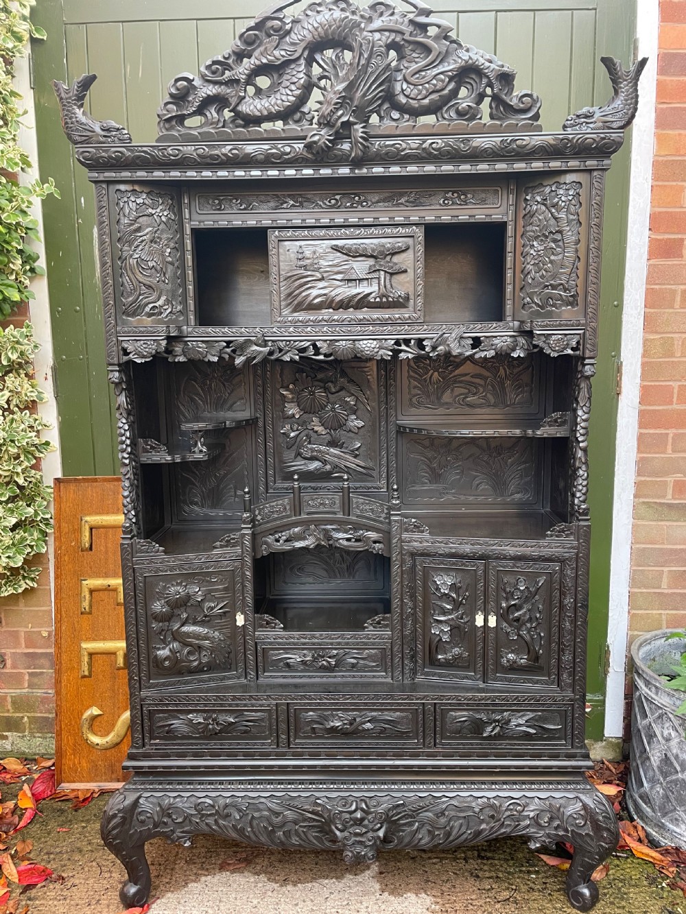 An early 20th-century Japanese ebonised timber shodana display cabinet featuring detailed dragon and floral relief carvings, asymmetrical shelving, small cupboard doors, and a carved stand base. View II