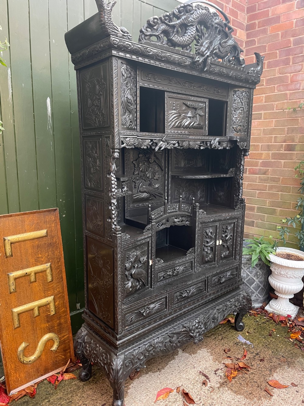 An early 20th-century Japanese ebonised timber shodana display cabinet featuring detailed dragon and floral relief carvings, asymmetrical shelving, small cupboard doors, and a carved stand base. View VI