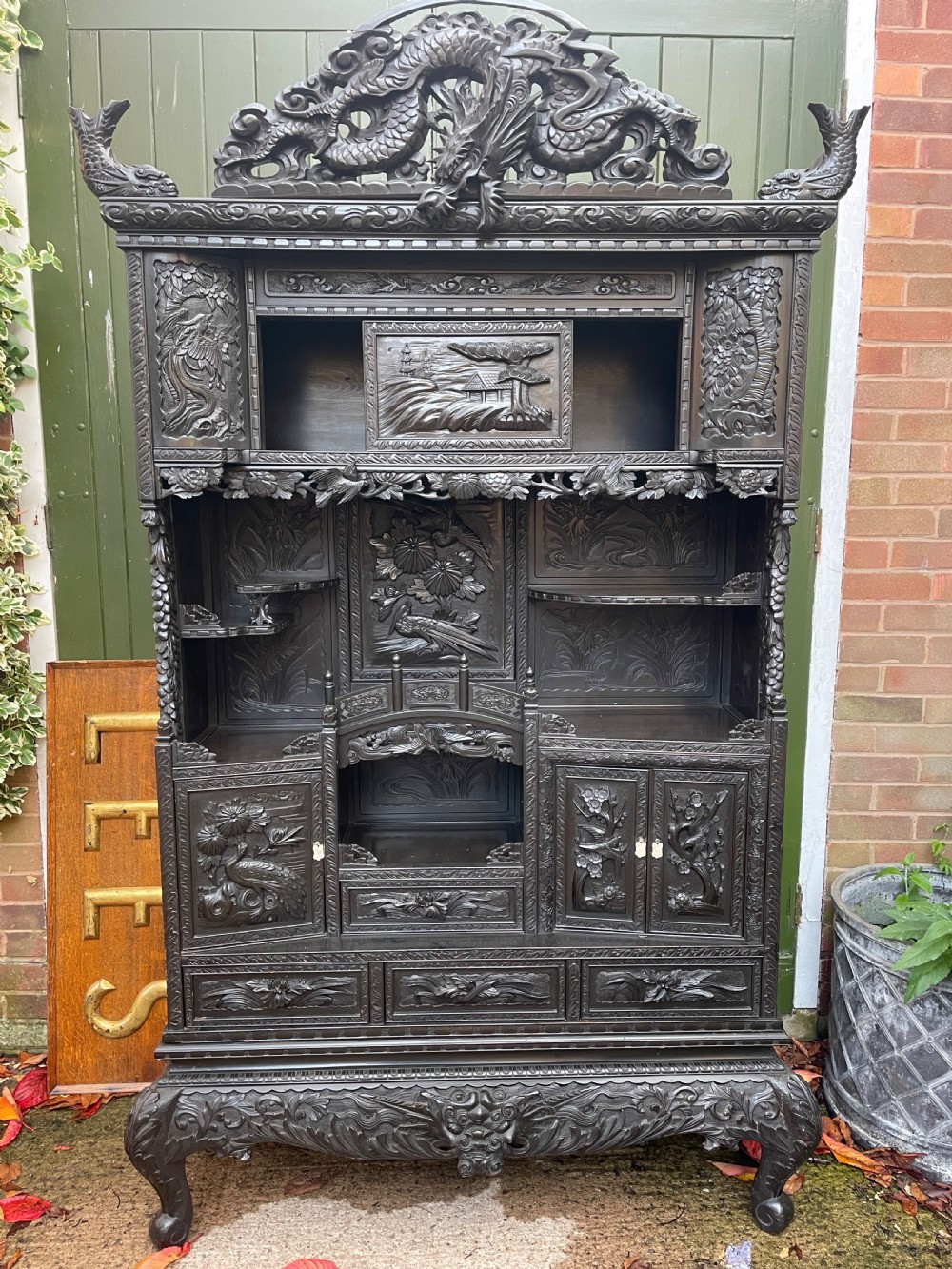 An early 20th-century Japanese ebonised timber shodana display cabinet featuring detailed dragon and floral relief carvings, asymmetrical shelving, small cupboard doors, and a carved stand base. View XII