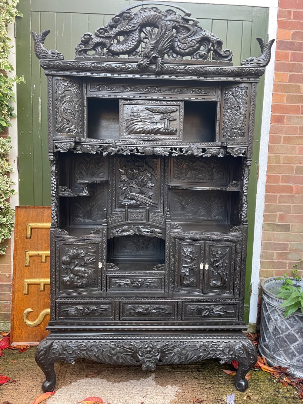 An early 20th-century Japanese ebonised timber shodana display cabinet featuring detailed dragon and floral relief carvings, asymmetrical shelving, small cupboard doors, and a carved stand base. View XIV