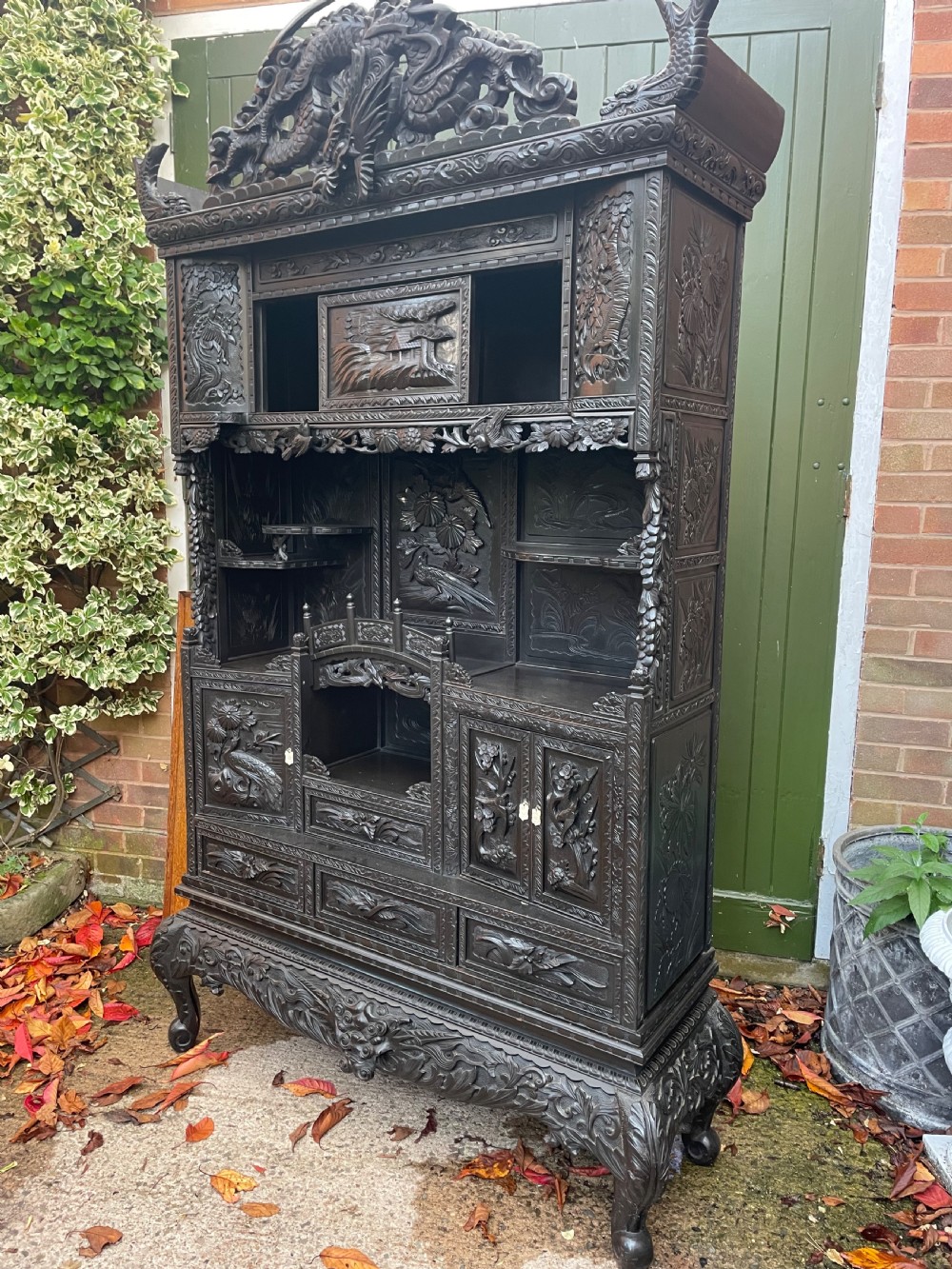 An early 20th-century Japanese ebonised timber shodana display cabinet featuring detailed dragon and floral relief carvings, asymmetrical shelving, small cupboard doors, and a carved stand base. View XVI