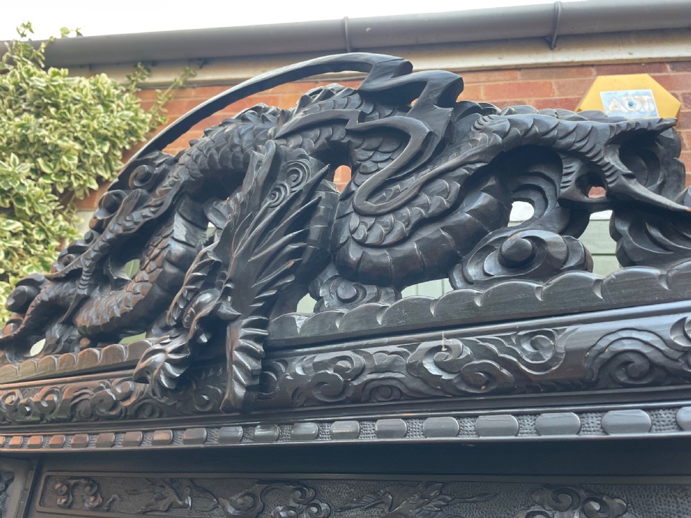 An early 20th-century Japanese ebonised timber shodana display cabinet featuring detailed dragon and floral relief carvings, asymmetrical shelving, small cupboard doors, and a carved stand base. View XIX