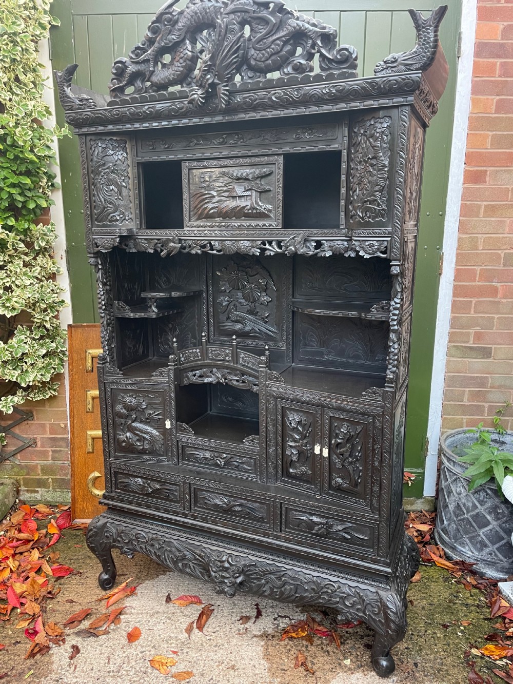 An early 20th-century Japanese ebonised timber shodana display cabinet featuring detailed dragon and floral relief carvings, asymmetrical shelving, small cupboard doors, and a carved stand base. View XX