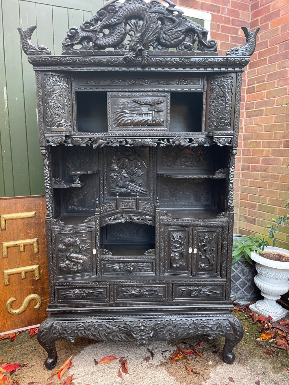 An early 20th-century Japanese ebonised timber shodana display cabinet featuring detailed dragon and floral relief carvings, asymmetrical shelving, small cupboard doors, and a carved stand base. View XXI