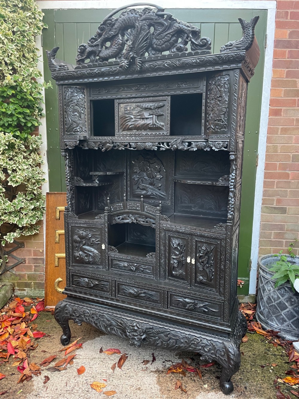 An early 20th-century Japanese ebonised timber shodana display cabinet featuring detailed dragon and floral relief carvings, asymmetrical shelving, small cupboard doors, and a carved stand base. View XXII