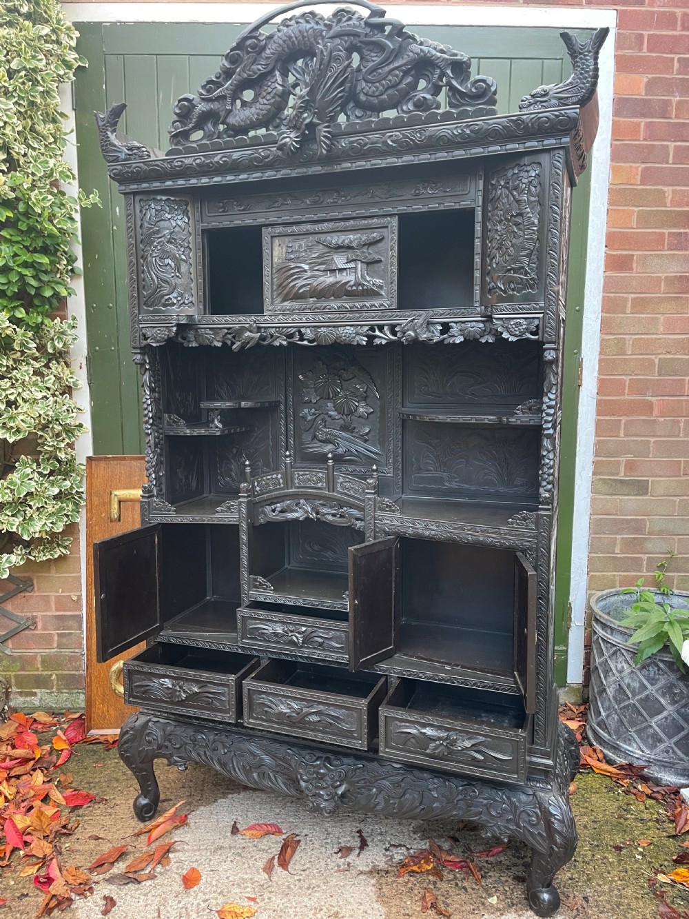 An early 20th-century Japanese ebonised timber shodana display cabinet featuring detailed dragon and floral relief carvings, asymmetrical shelving, small cupboard doors, and a carved stand base. View XXIV