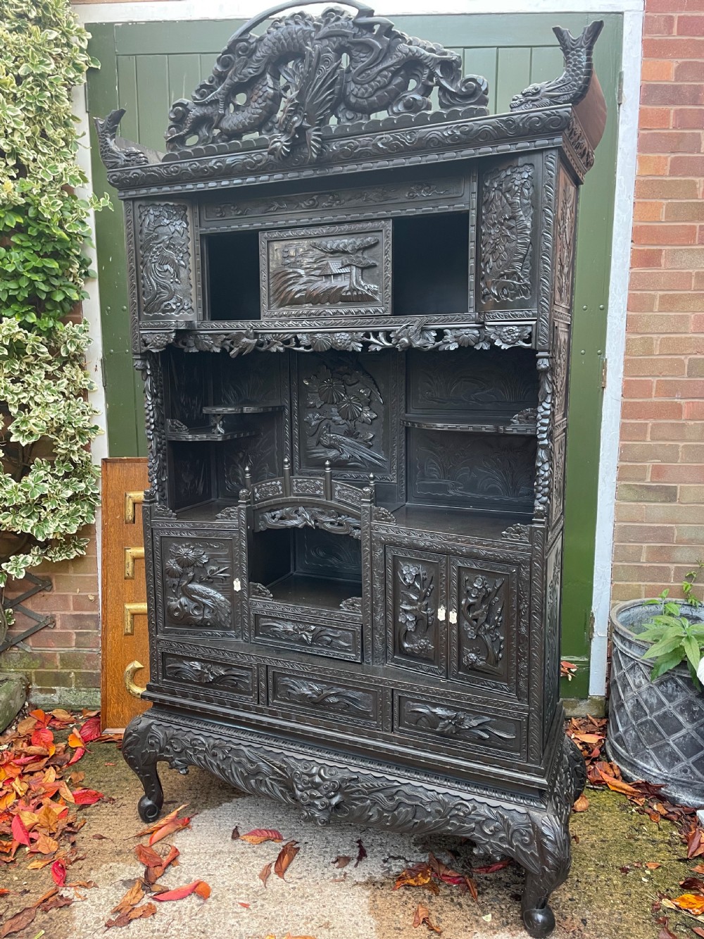 An early 20th-century Japanese ebonised timber shodana display cabinet featuring detailed dragon and floral relief carvings, asymmetrical shelving, small cupboard doors, and a carved stand base. View XXV