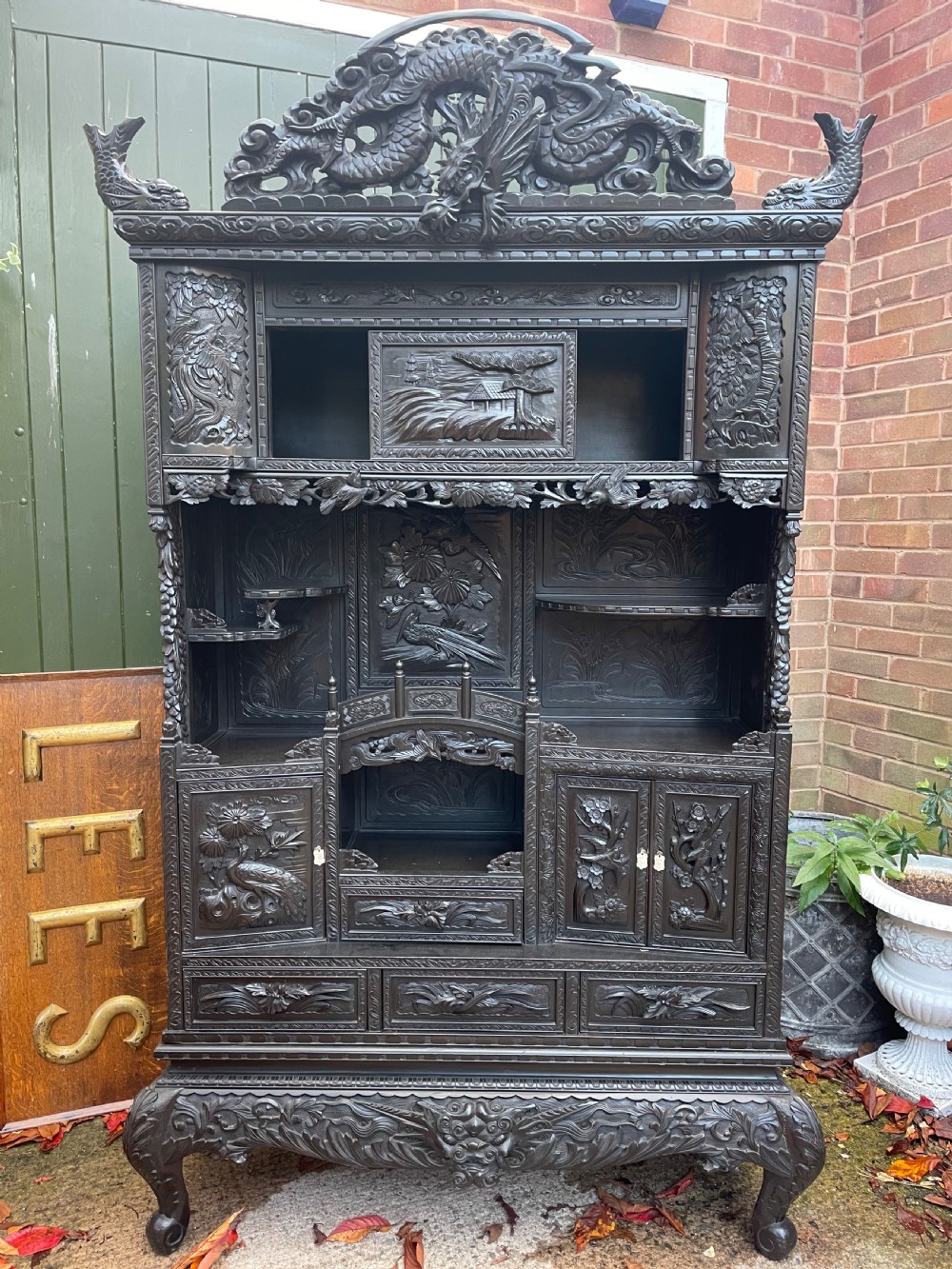 An early 20th-century Japanese ebonised timber shodana display cabinet featuring detailed dragon and floral relief carvings, asymmetrical shelving, small cupboard doors, and a carved stand base. View XXVI