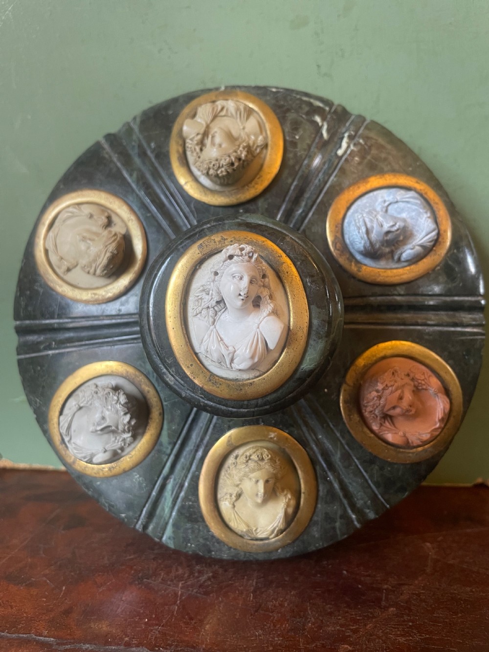 A circular dark green carved Prato Verde marble inkstand with fluted sides, set with six oval carved Vesuvian lava portrait cameos framed in brass mounts around a central raised finial. View VIII