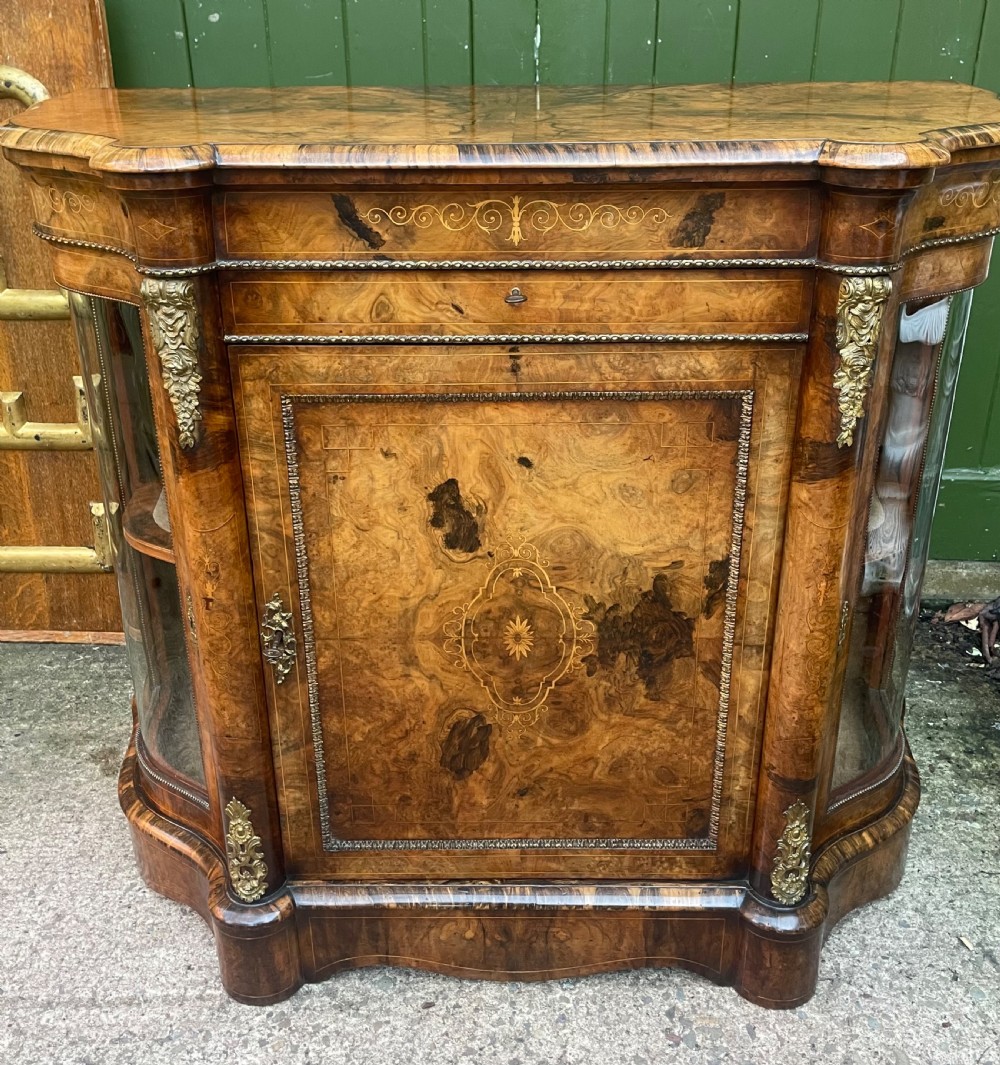 A mid-19th century Victorian burr walnut credenza featuring boxwood marquetry inlay, gilded ormolu mounts, a central drawer, and curved glass side display doors.