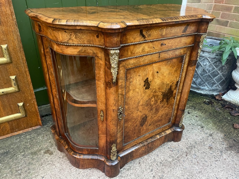 A mid-19th century Victorian burr walnut credenza featuring boxwood marquetry inlay, gilded ormolu mounts, a central drawer, and curved glass side display doors. View V