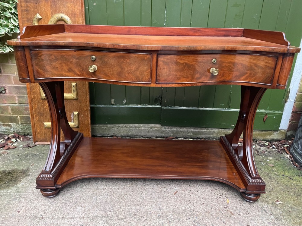 An early nineteenth-century mahogany serpentine side table featuring two drawers, a gallery top, curved end supports, and a lower shelf.