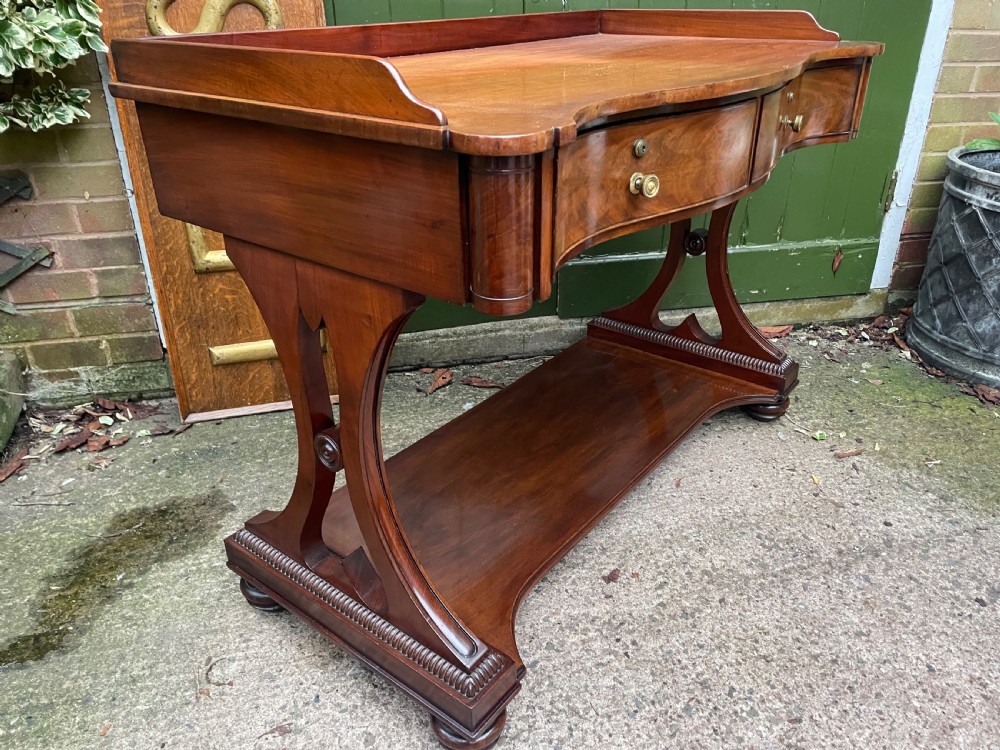 An early nineteenth-century mahogany serpentine side table featuring two drawers, a gallery top, curved end supports, and a lower shelf. View IX