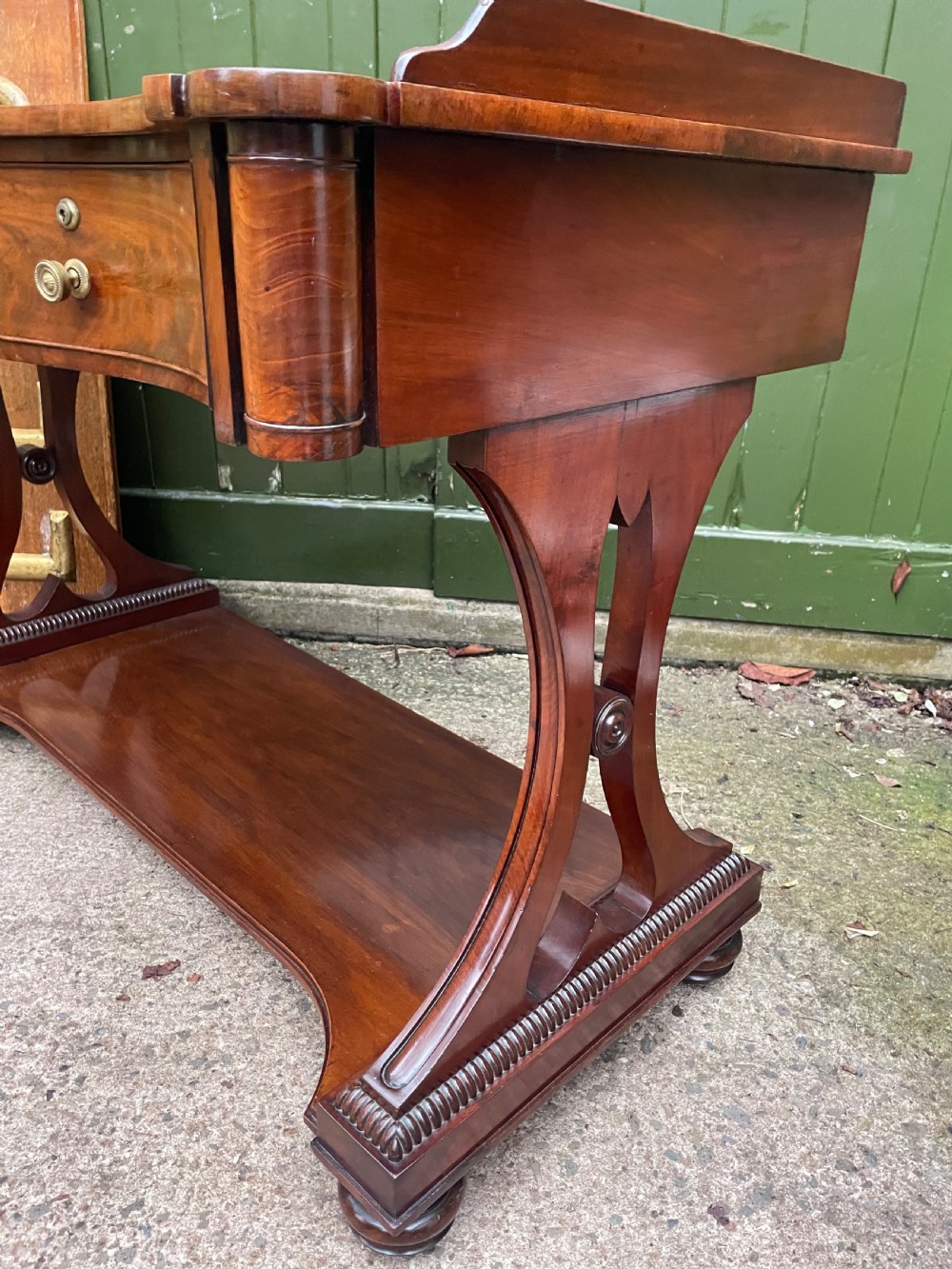 An early nineteenth-century mahogany serpentine side table featuring two drawers, a gallery top, curved end supports, and a lower shelf. View XII