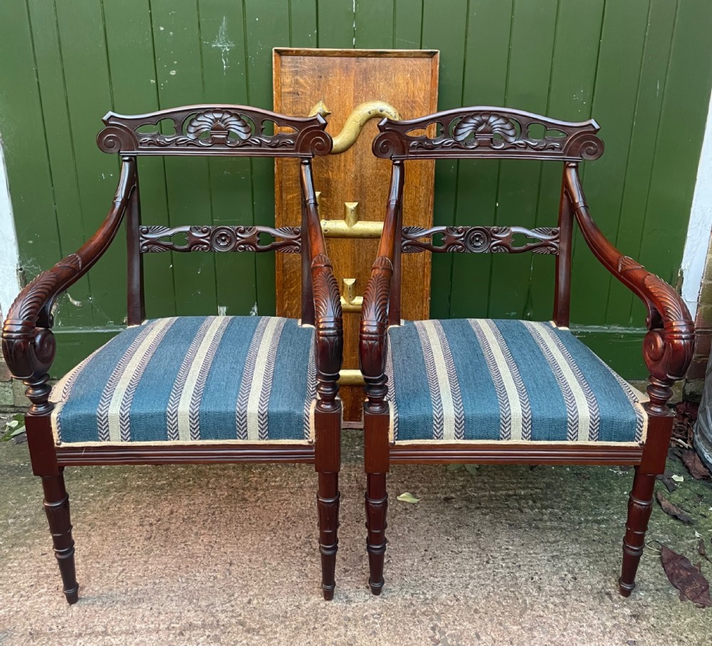 A pair of early 19th-century carved mahogany salon armchairs featuring carved crest rails, scrolled armrests, turned front legs, and blue striped fabric upholstery.