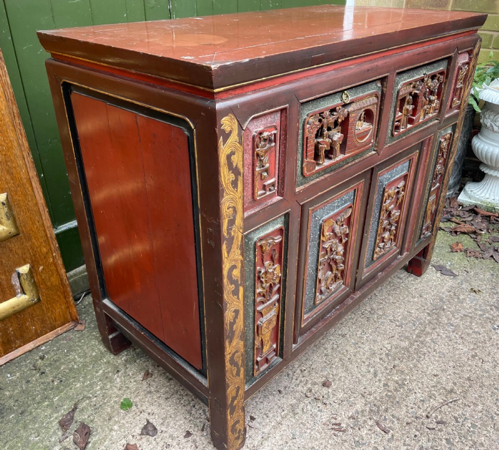 A late nineteenth-century Chinese Qing Dynasty red lacquered side cabinet featuring relief-carved gilded wood panels depicted with architectural scenes, figures, and foliage. View V