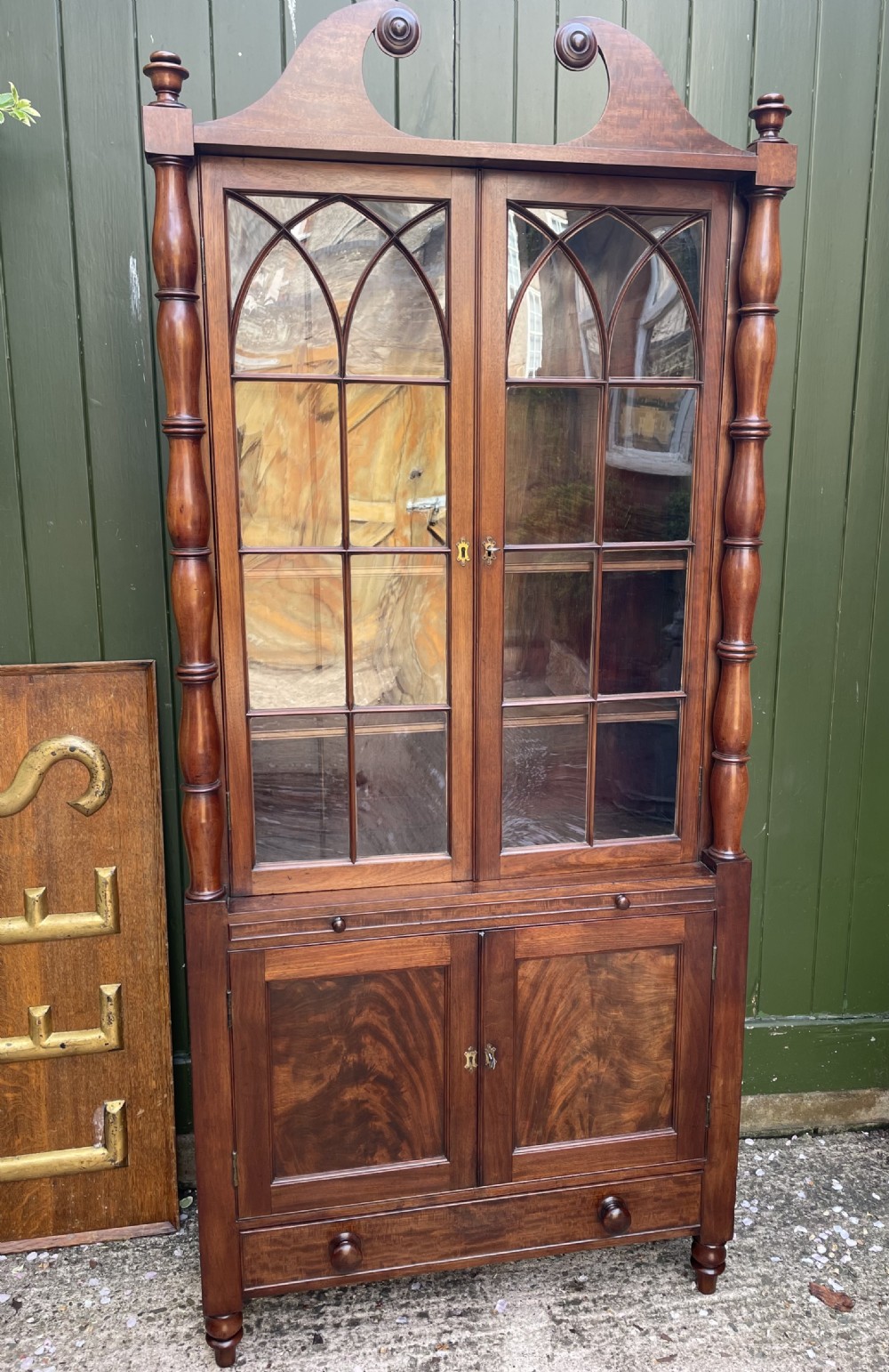 An early nineteenth-century Regency mahogany glazed bookcase cabinet featuring a scroll pediment, Gothic arch astragal glazing, flame mahogany lower cupboard panels, and turned side columns. View II