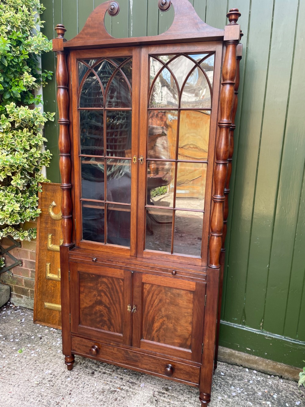 An early nineteenth-century Regency mahogany glazed bookcase cabinet featuring a scroll pediment, Gothic arch astragal glazing, flame mahogany lower cupboard panels, and turned side columns. View III