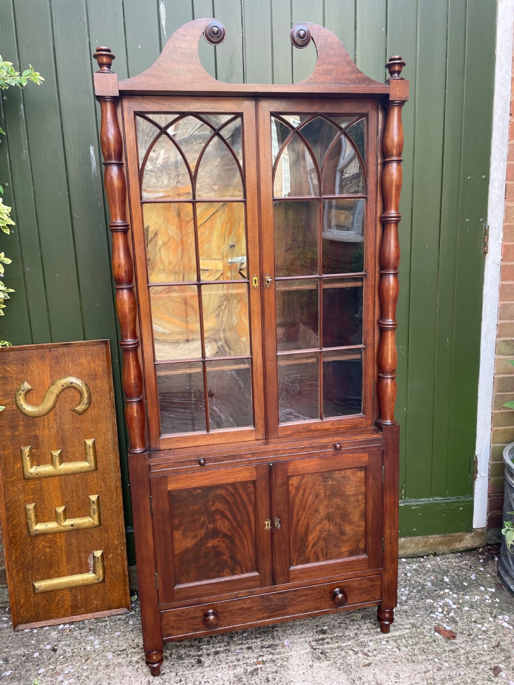 An early nineteenth-century Regency mahogany glazed bookcase cabinet featuring a scroll pediment, Gothic arch astragal glazing, flame mahogany lower cupboard panels, and turned side columns. View V