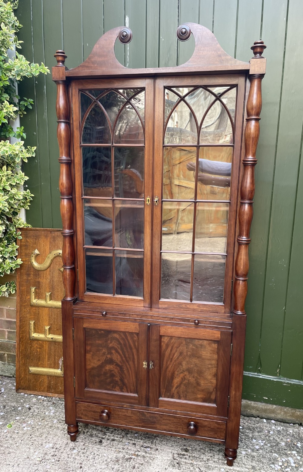 An early nineteenth-century Regency mahogany glazed bookcase cabinet featuring a scroll pediment, Gothic arch astragal glazing, flame mahogany lower cupboard panels, and turned side columns. View VIII