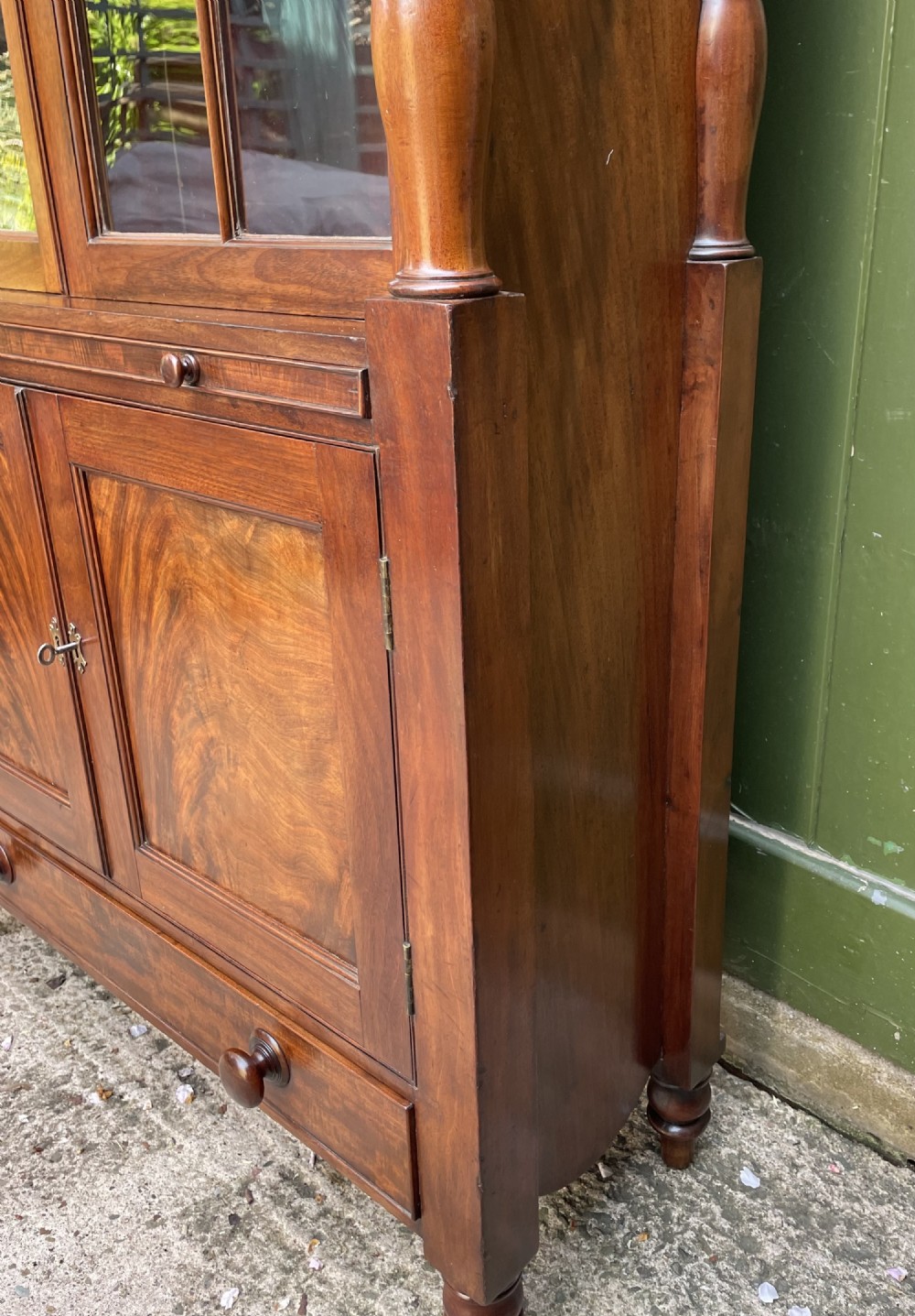 An early nineteenth-century Regency mahogany glazed bookcase cabinet featuring a scroll pediment, Gothic arch astragal glazing, flame mahogany lower cupboard panels, and turned side columns. View XI