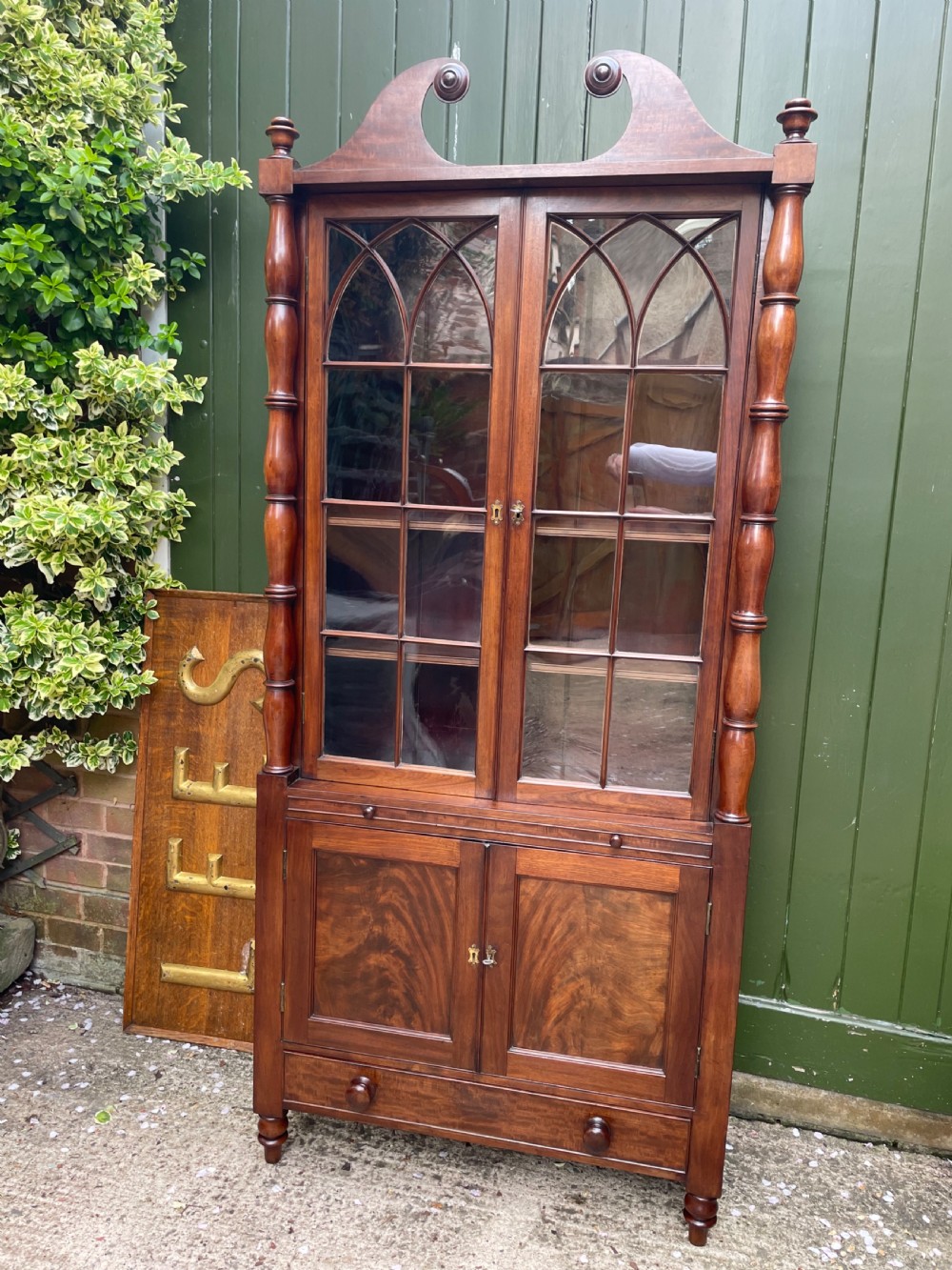 An early nineteenth-century Regency mahogany glazed bookcase cabinet featuring a scroll pediment, Gothic arch astragal glazing, flame mahogany lower cupboard panels, and turned side columns. View XIII