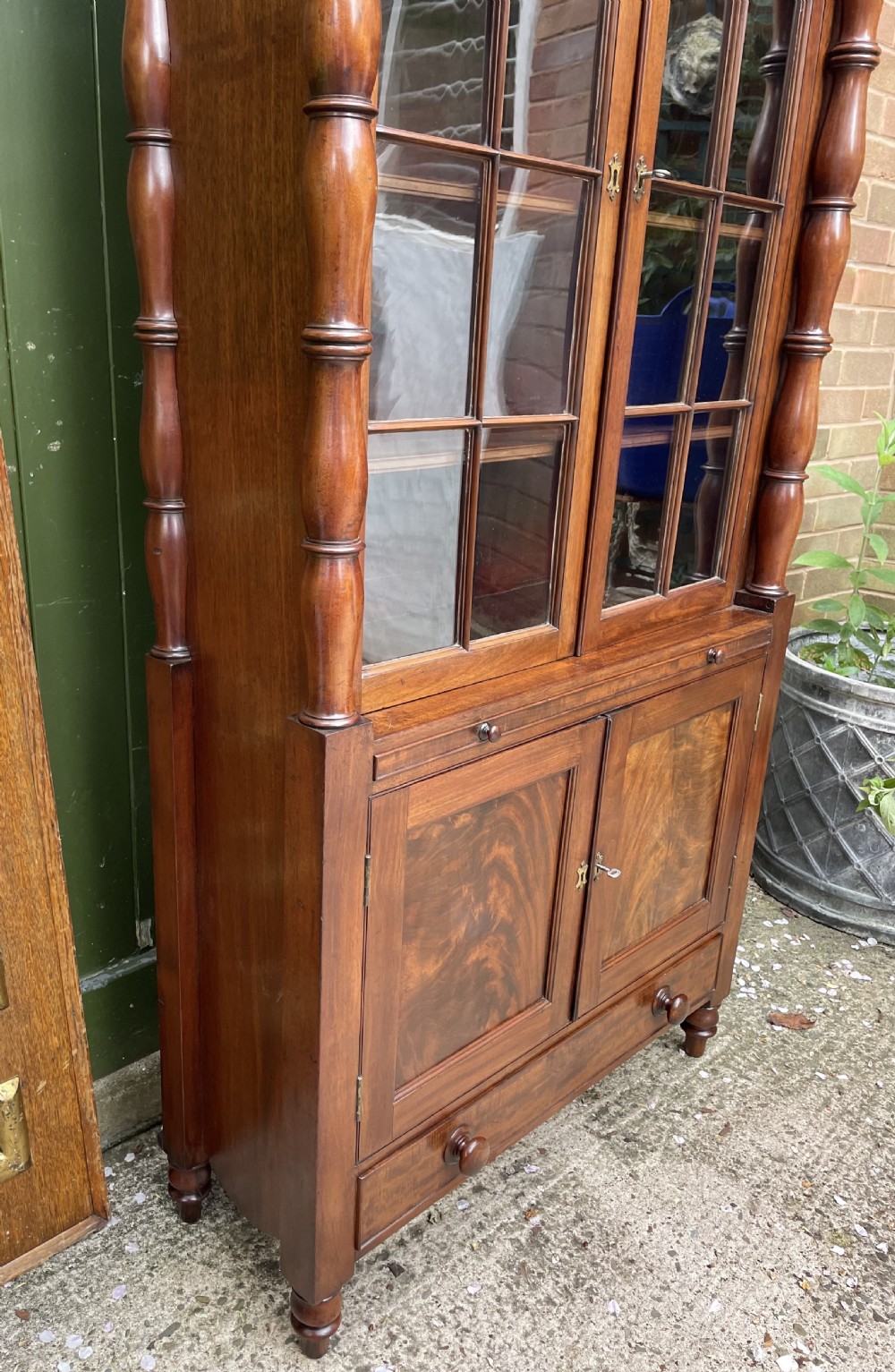An early nineteenth-century Regency mahogany glazed bookcase cabinet featuring a scroll pediment, Gothic arch astragal glazing, flame mahogany lower cupboard panels, and turned side columns. View XIV
