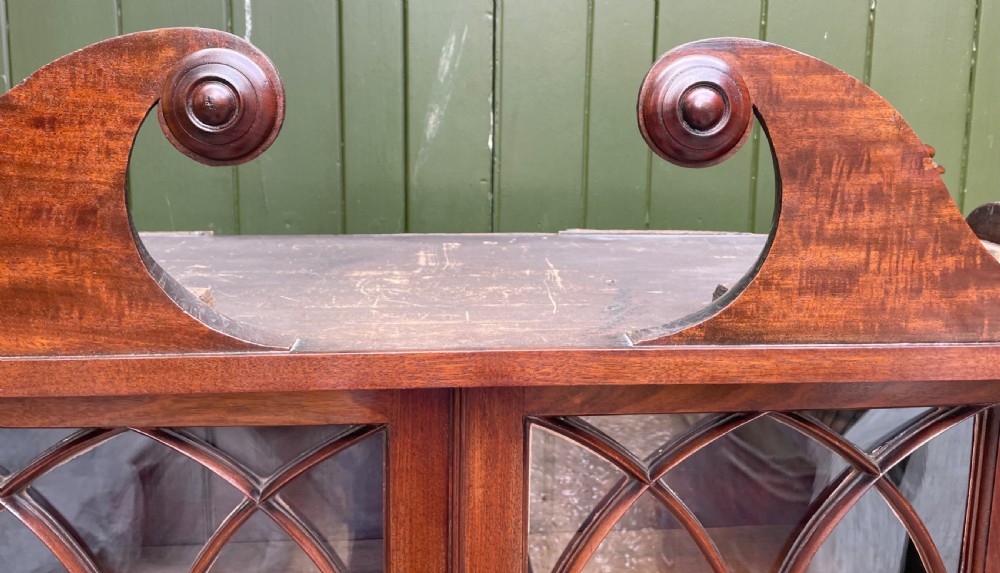 An early nineteenth-century Regency mahogany glazed bookcase cabinet featuring a scroll pediment, Gothic arch astragal glazing, flame mahogany lower cupboard panels, and turned side columns. View XVI