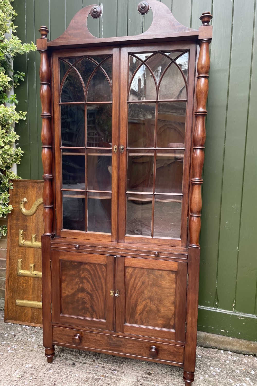 An early nineteenth-century Regency mahogany glazed bookcase cabinet featuring a scroll pediment, Gothic arch astragal glazing, flame mahogany lower cupboard panels, and turned side columns. View XVIII