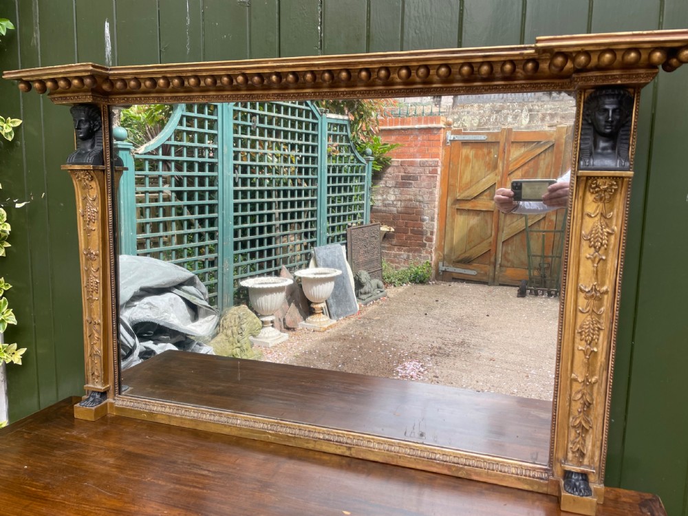 An early nineteenth-century Regency giltwood overmantel mirror featuring ball moulding along the cornice and ebonised Egyptian sphinx heads atop decorative pilasters.