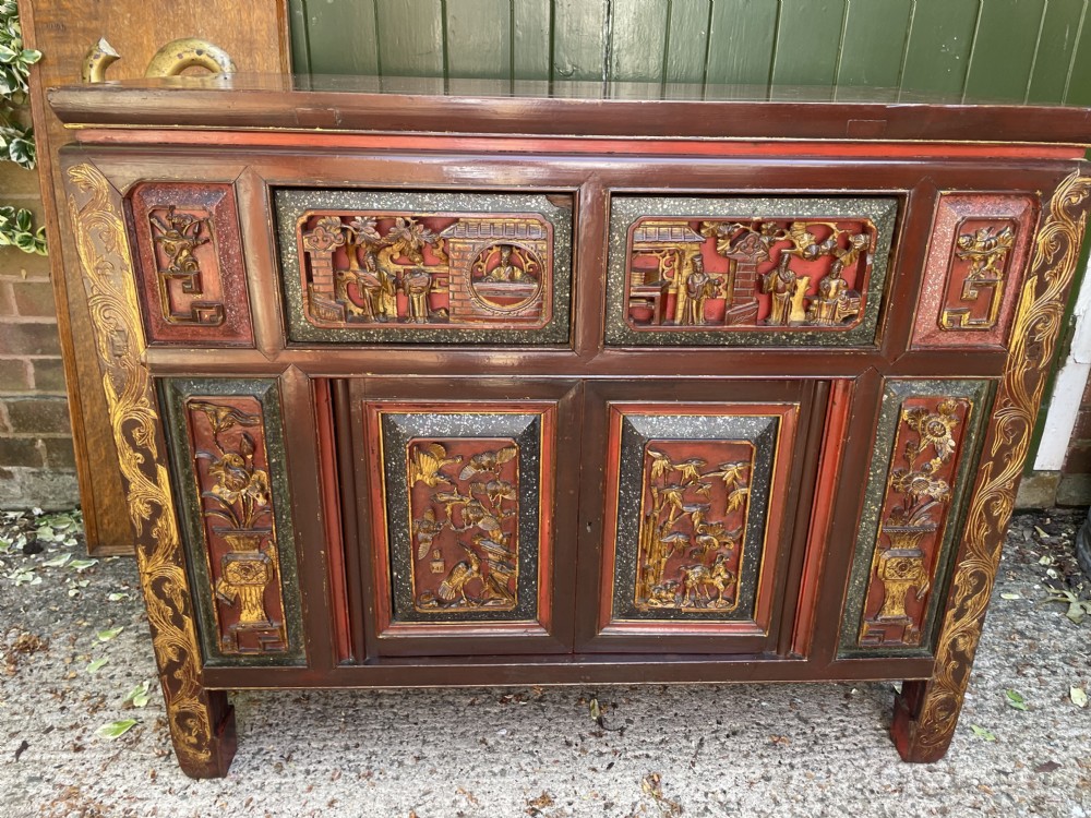 A late nineteenth-century Chinese Qing Dynasty red lacquered side cabinet featuring relief-carved gilded wood panels depicted with architectural scenes, figures, and foliage.