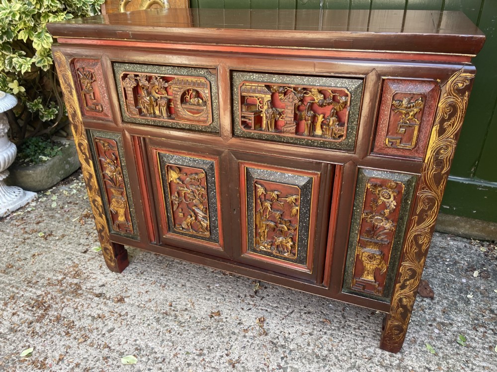 A late nineteenth-century Chinese Qing Dynasty red lacquered side cabinet featuring relief-carved gilded wood panels depicted with architectural scenes, figures, and foliage. View II