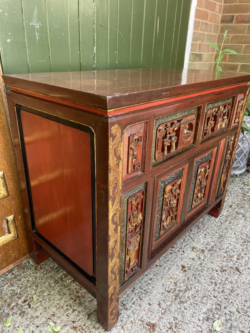 A late nineteenth-century Chinese Qing Dynasty red lacquered side cabinet featuring relief-carved gilded wood panels depicted with architectural scenes, figures, and foliage. View XX