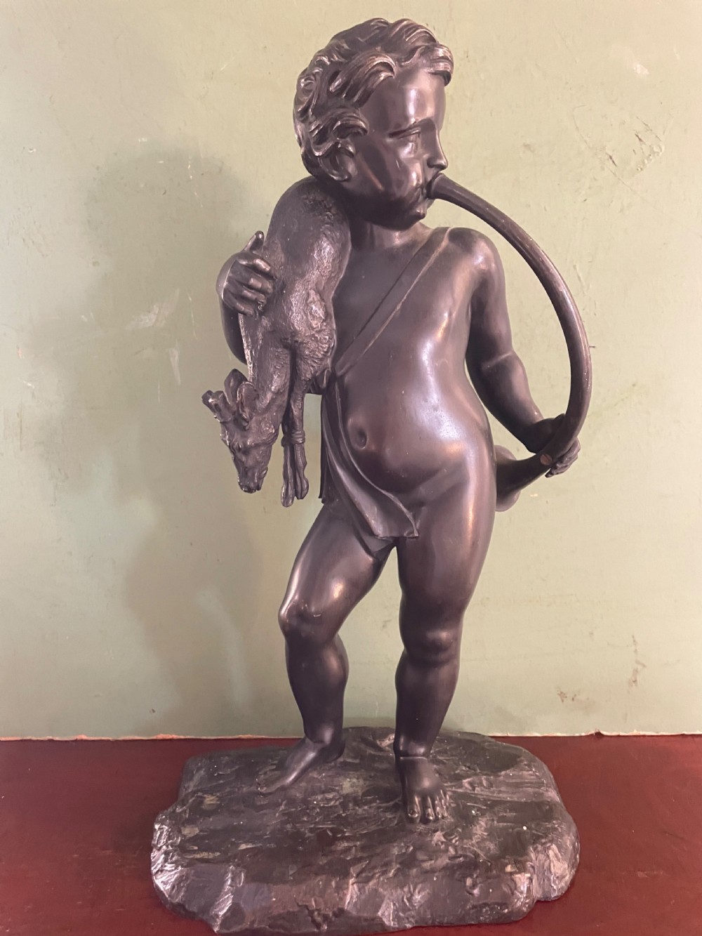 A dark patinated bronze sculpture depicting a standing putto hunter blowing a curved horn while slinging a small deer over one shoulder.