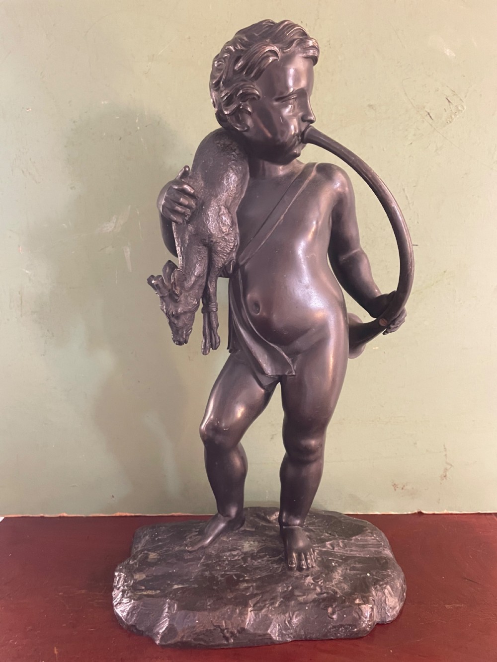 A dark patinated bronze sculpture depicting a standing putto hunter blowing a curved horn while slinging a small deer over one shoulder. View II