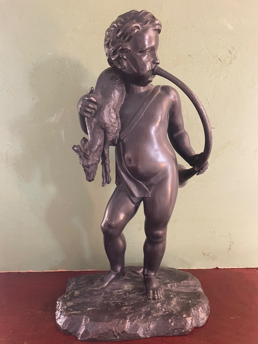 A dark patinated bronze sculpture depicting a standing putto hunter blowing a curved horn while slinging a small deer over one shoulder. View III