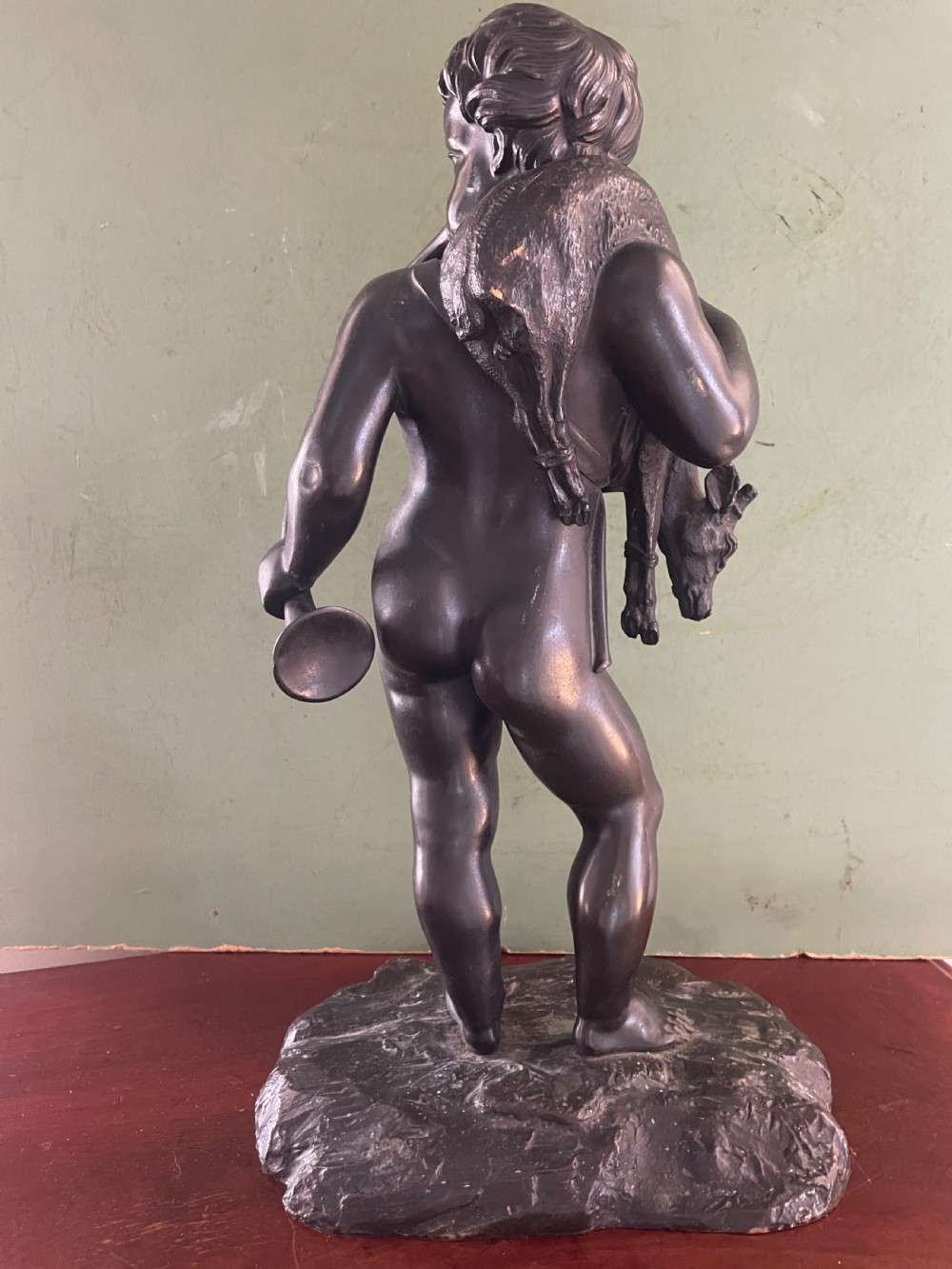 A dark patinated bronze sculpture depicting a standing putto hunter blowing a curved horn while slinging a small deer over one shoulder. View V