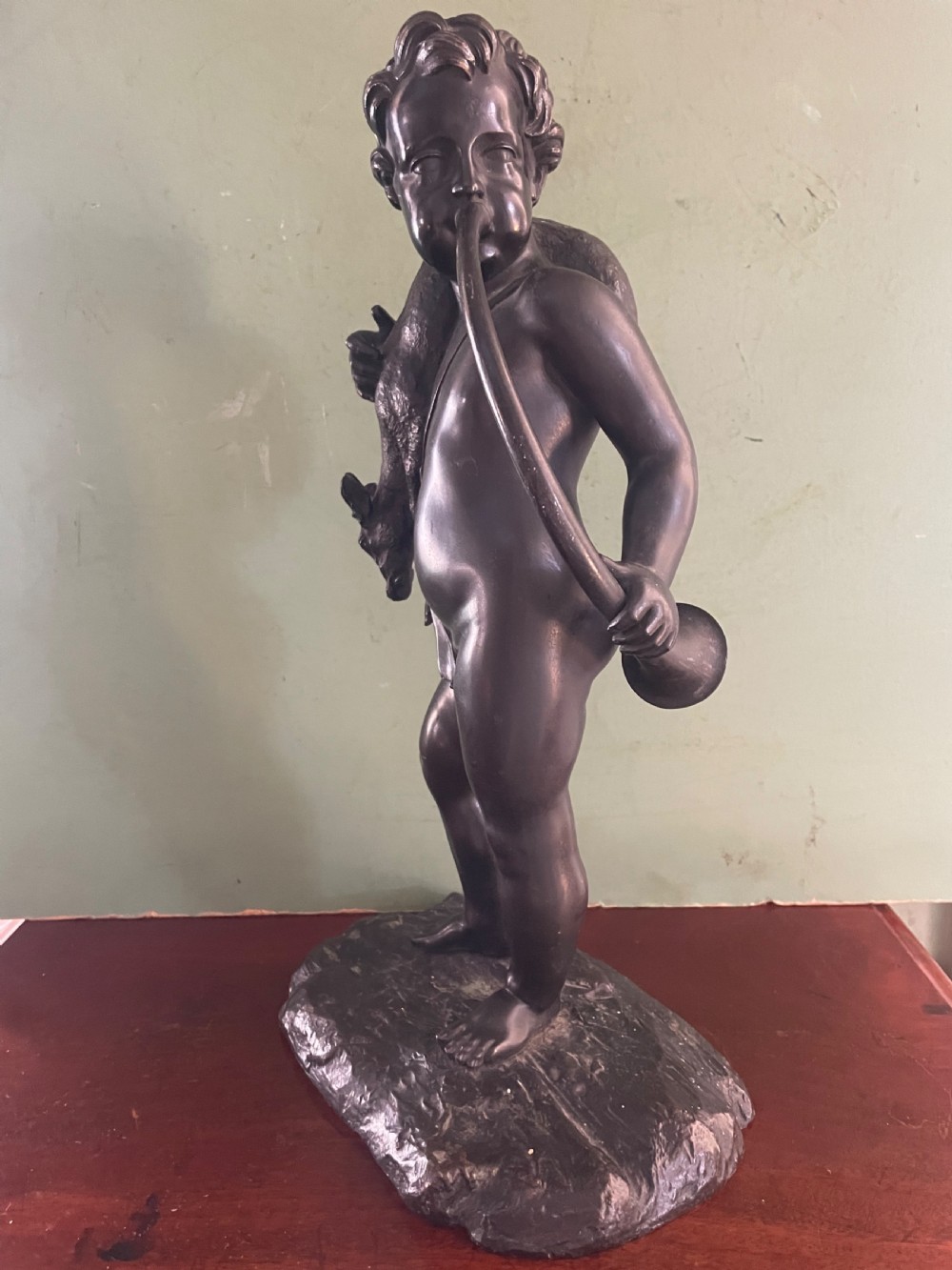 A dark patinated bronze sculpture depicting a standing putto hunter blowing a curved horn while slinging a small deer over one shoulder. View VI