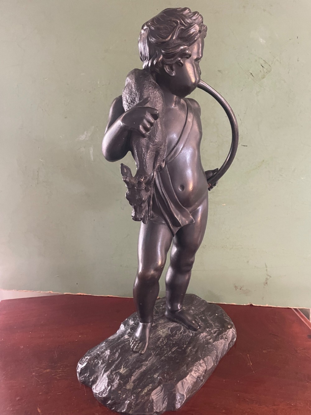 A dark patinated bronze sculpture depicting a standing putto hunter blowing a curved horn while slinging a small deer over one shoulder. View VIII