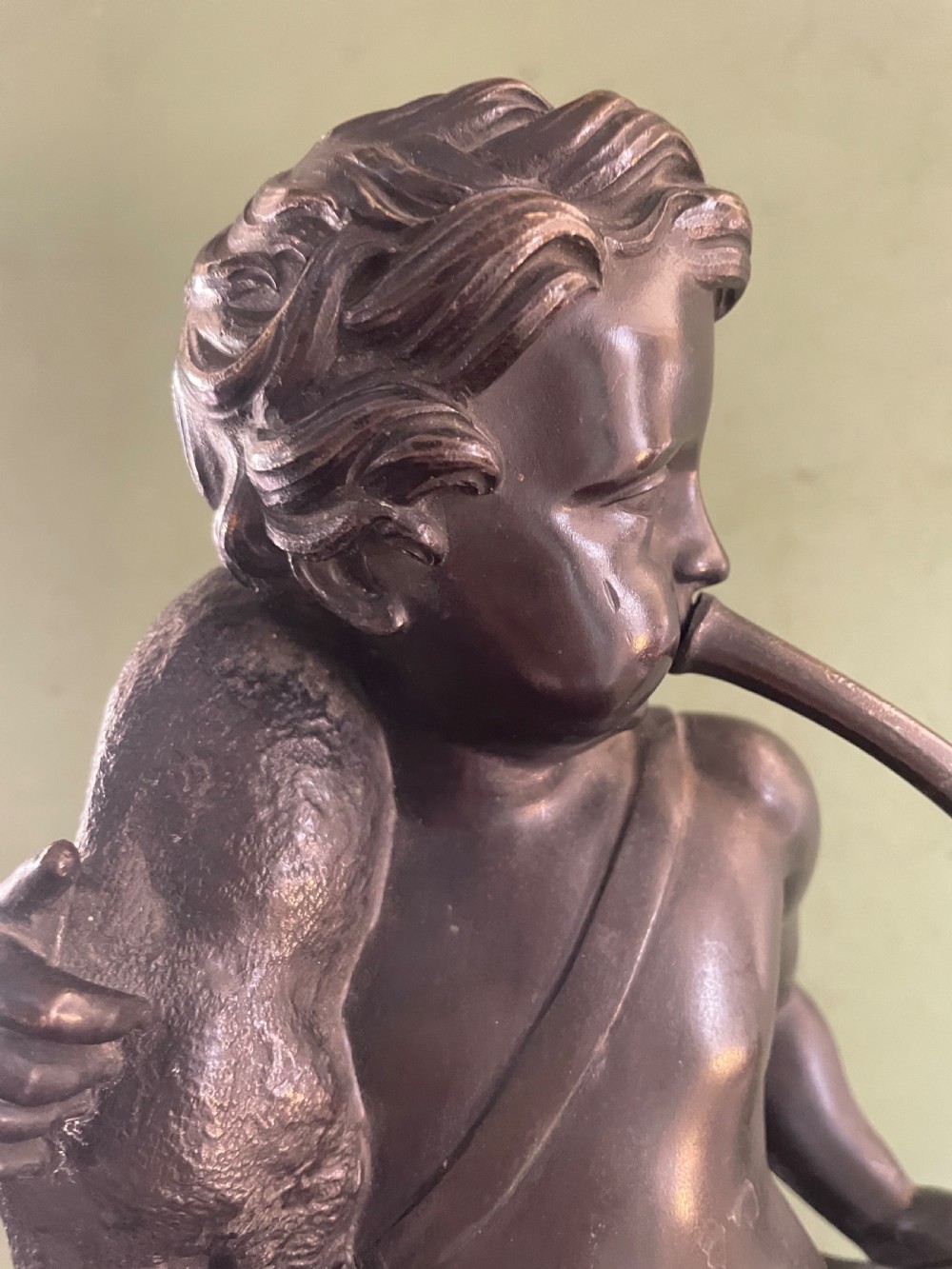 A dark patinated bronze sculpture depicting a standing putto hunter blowing a curved horn while slinging a small deer over one shoulder. View IX