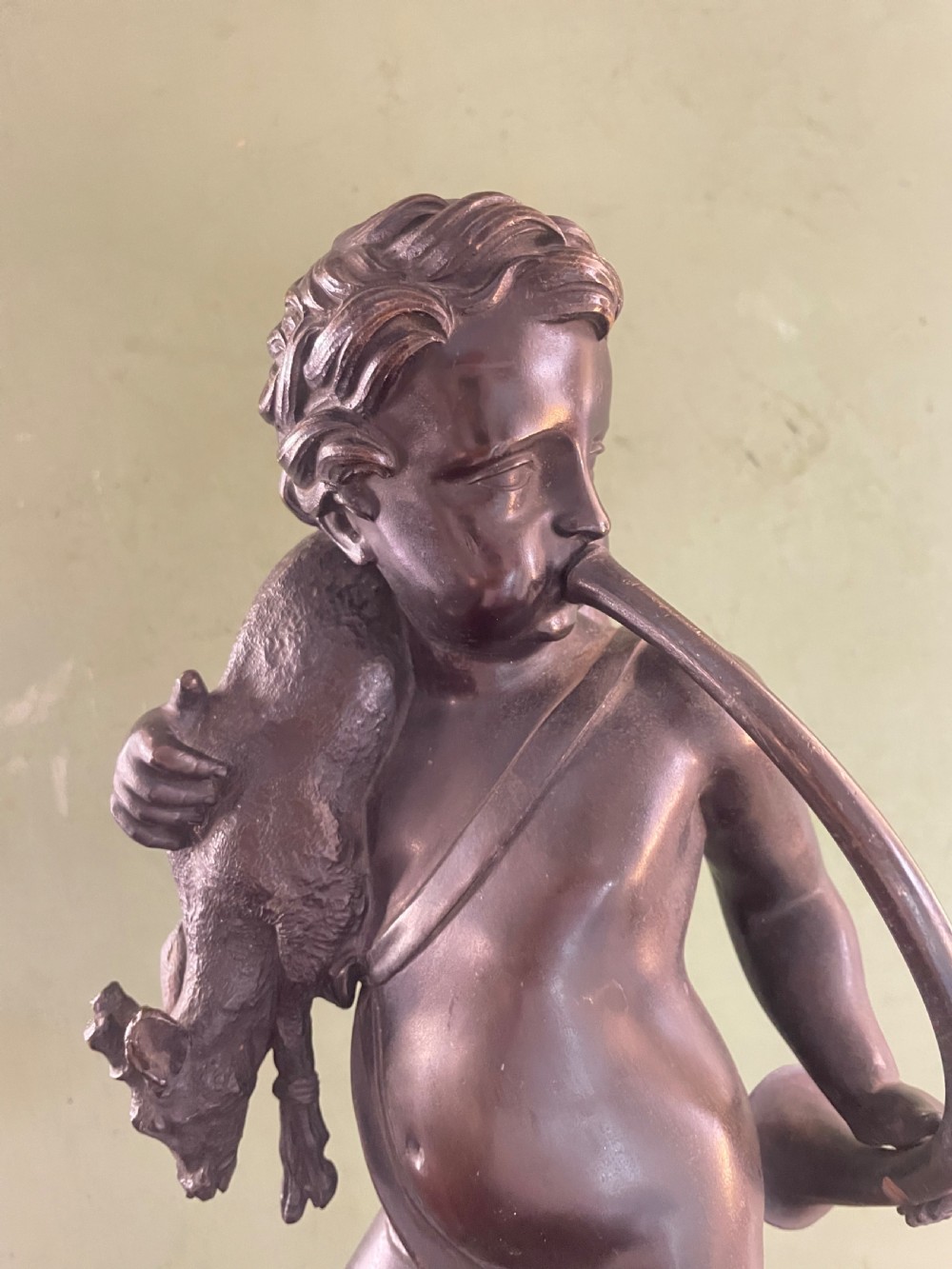 A dark patinated bronze sculpture depicting a standing putto hunter blowing a curved horn while slinging a small deer over one shoulder. View X