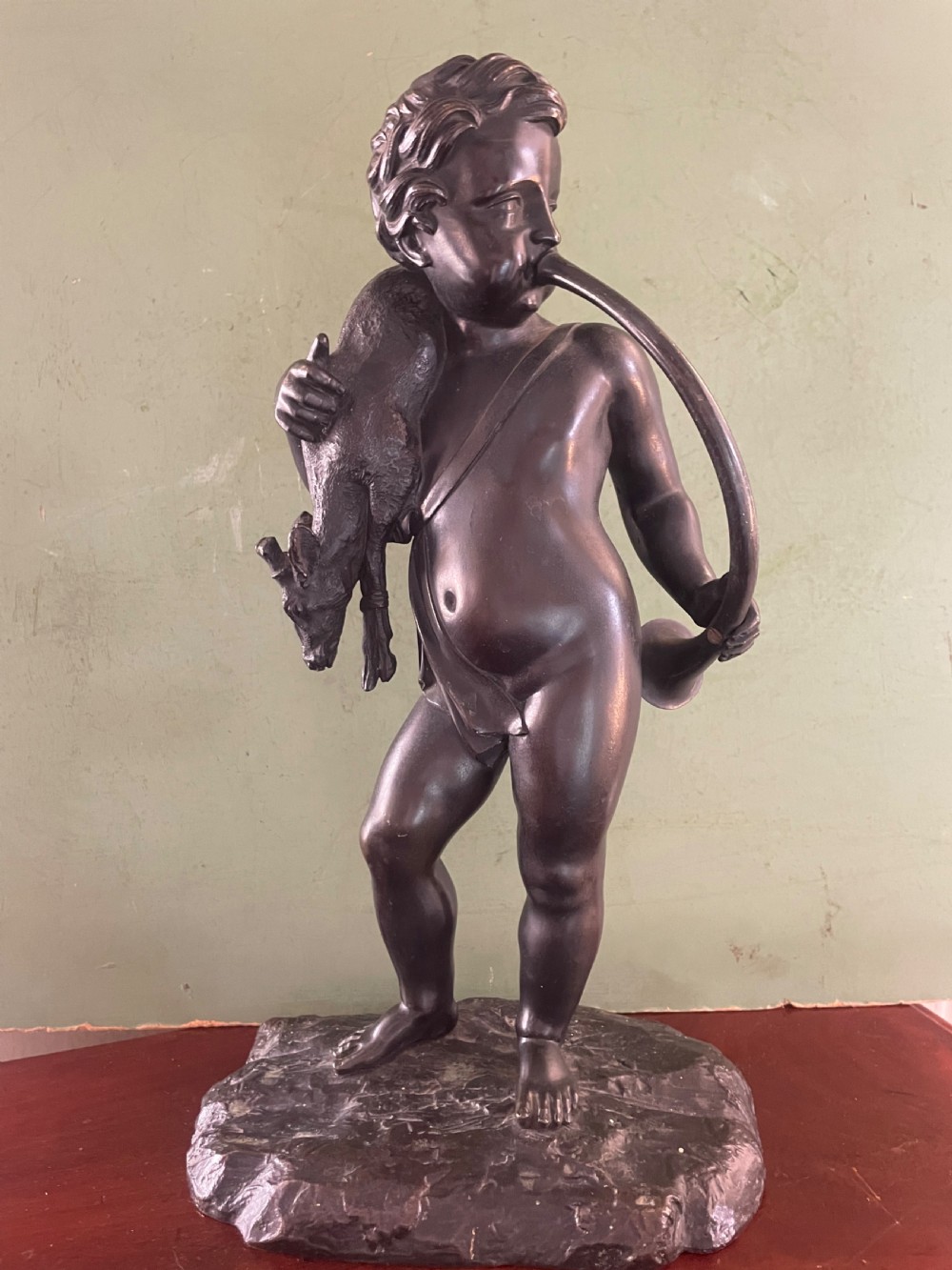 A dark patinated bronze sculpture depicting a standing putto hunter blowing a curved horn while slinging a small deer over one shoulder. View XII