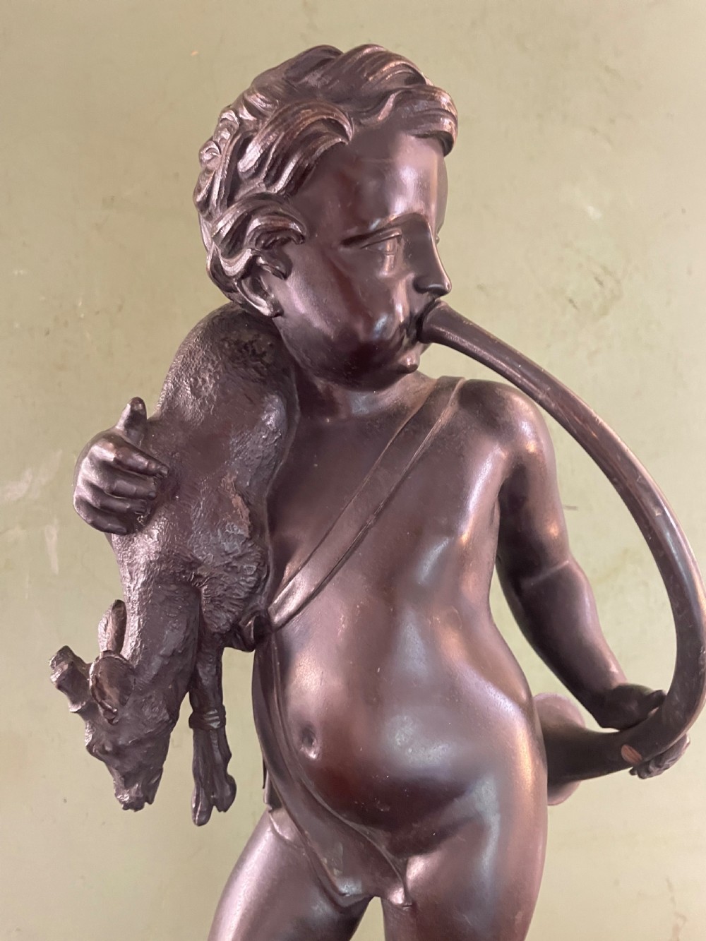 A dark patinated bronze sculpture depicting a standing putto hunter blowing a curved horn while slinging a small deer over one shoulder. View XIII