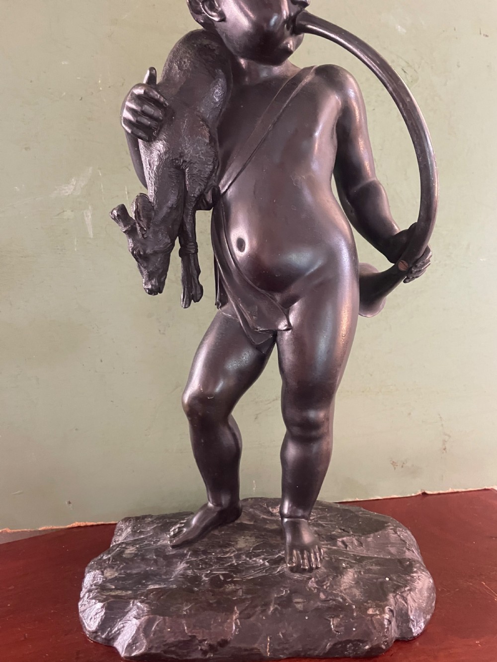 A dark patinated bronze sculpture depicting a standing putto hunter blowing a curved horn while slinging a small deer over one shoulder. View XIV