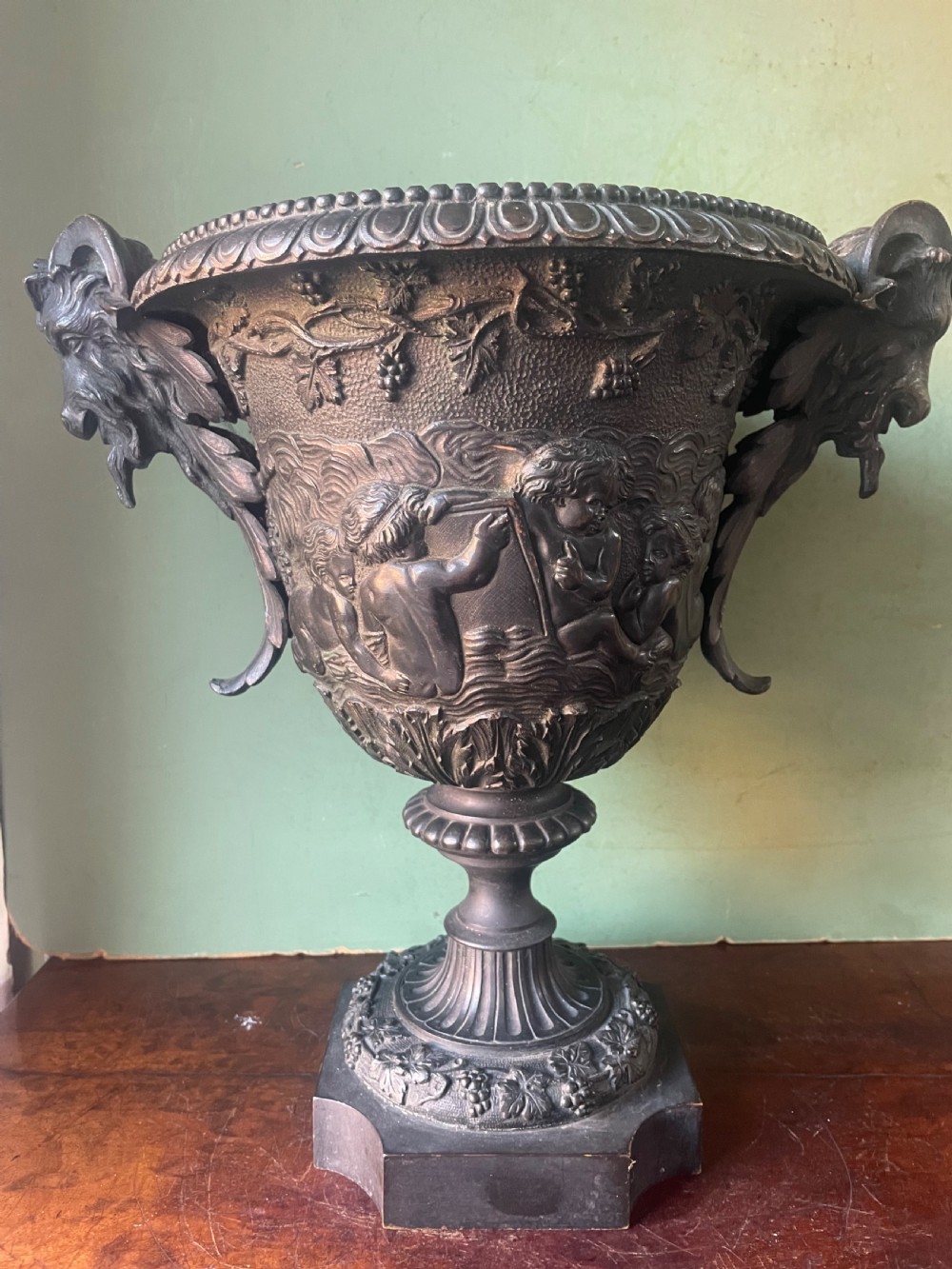 A 19th-century French dark bronze campana-shaped urn featuring relief carving of putti, satyr mask handles, and a square pedestal base. View V