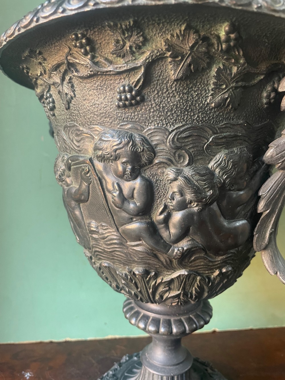 A 19th-century French dark bronze campana-shaped urn featuring relief carving of putti, satyr mask handles, and a square pedestal base. View VI