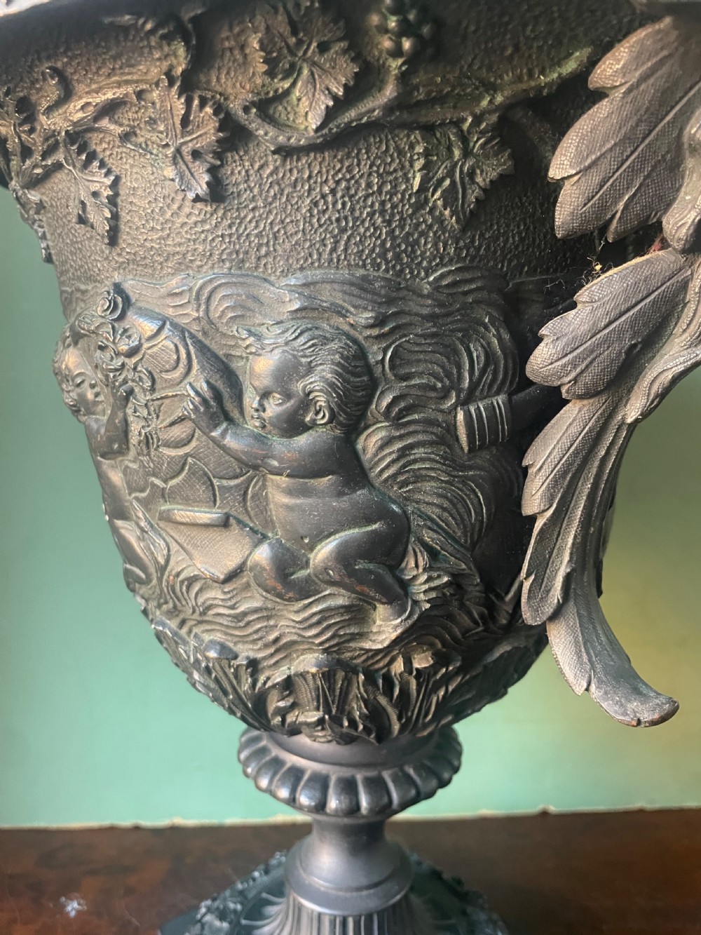 A 19th-century French dark bronze campana-shaped urn featuring relief carving of putti, satyr mask handles, and a square pedestal base. View XII