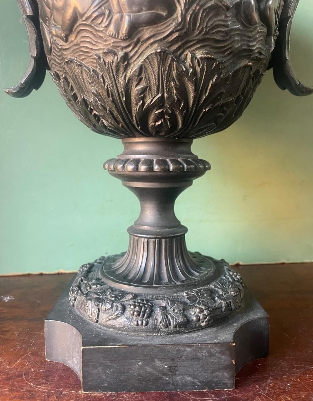 A 19th-century French dark bronze campana-shaped urn featuring relief carving of putti, satyr mask handles, and a square pedestal base. View XIII