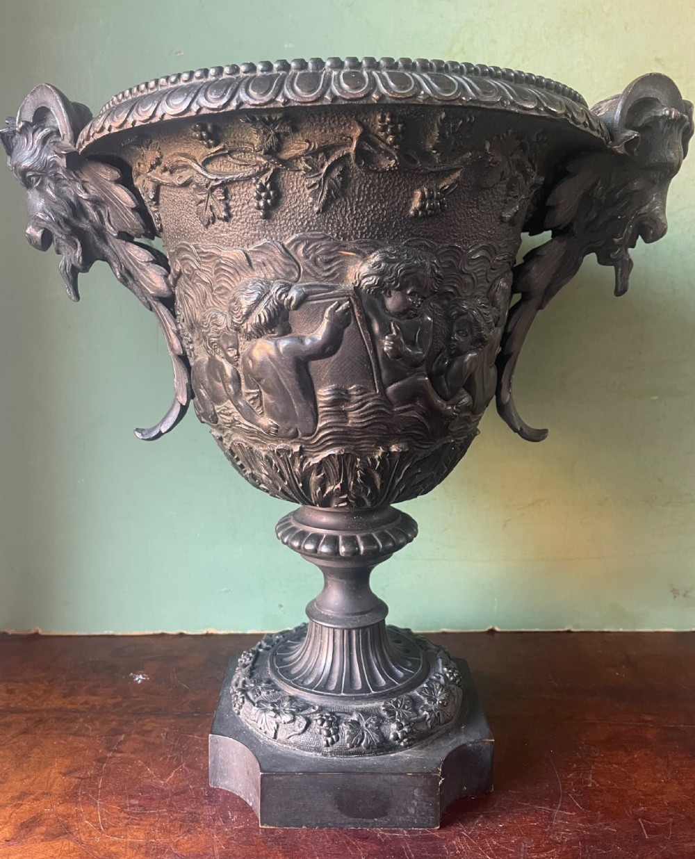 A 19th-century French dark bronze campana-shaped urn featuring relief carving of putti, satyr mask handles, and a square pedestal base. View XVI